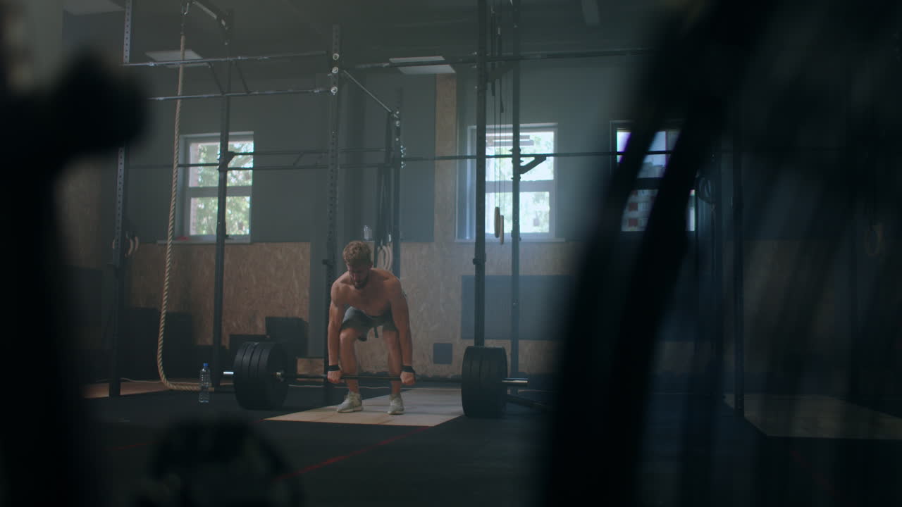 Wide shot of young Caucasian extreme weightlifting athlete man working out with heavy barbell in large hardcore gym hall.