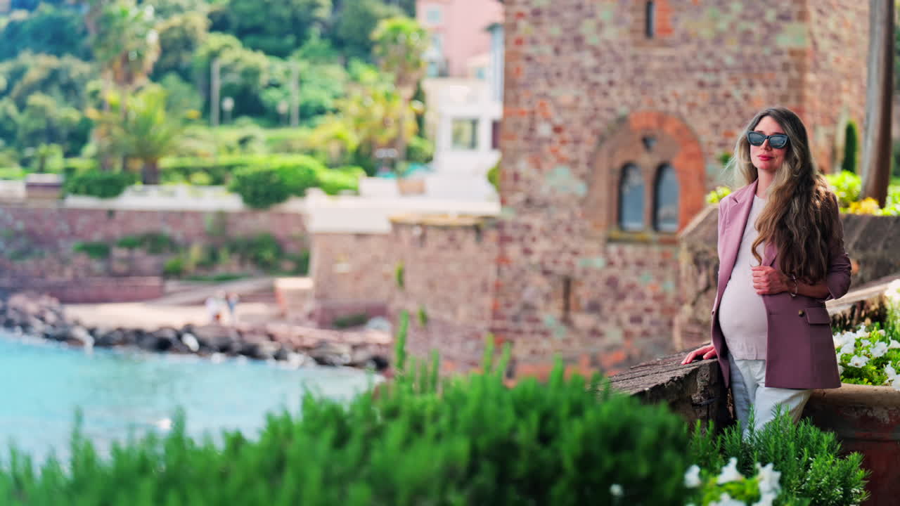 Pregnant woman with long hair, wearing sunglasses standing with the Chateau de la Napoule Castle in Mandelieu-La Napoule, France in the background