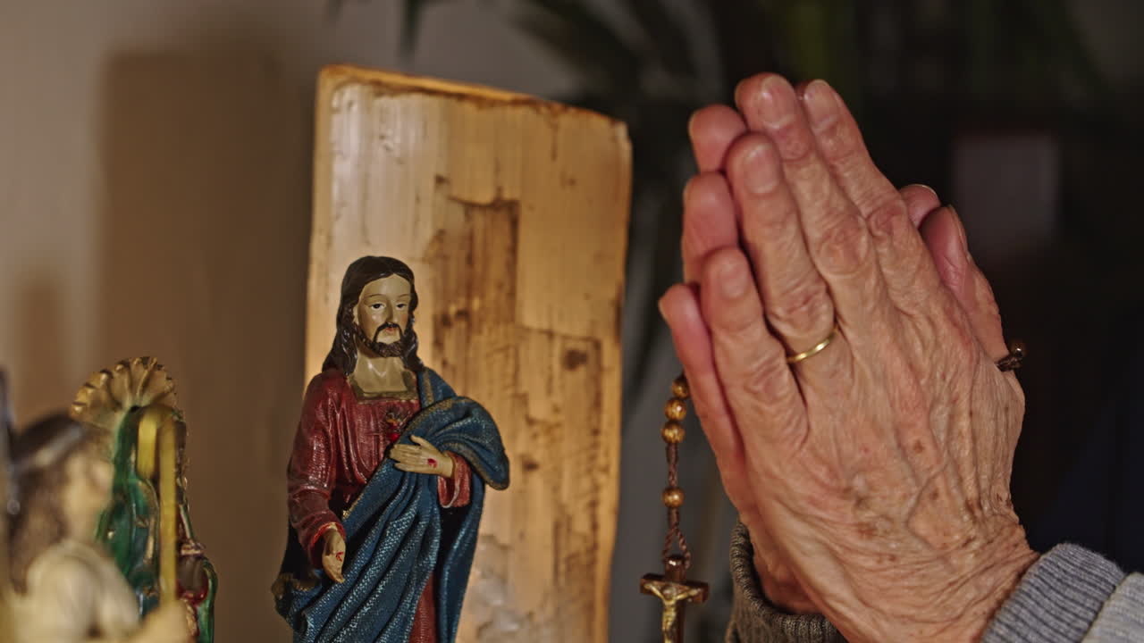 Primo piano di mani anziane in preghiera con rosario e statuetta di Gesù.