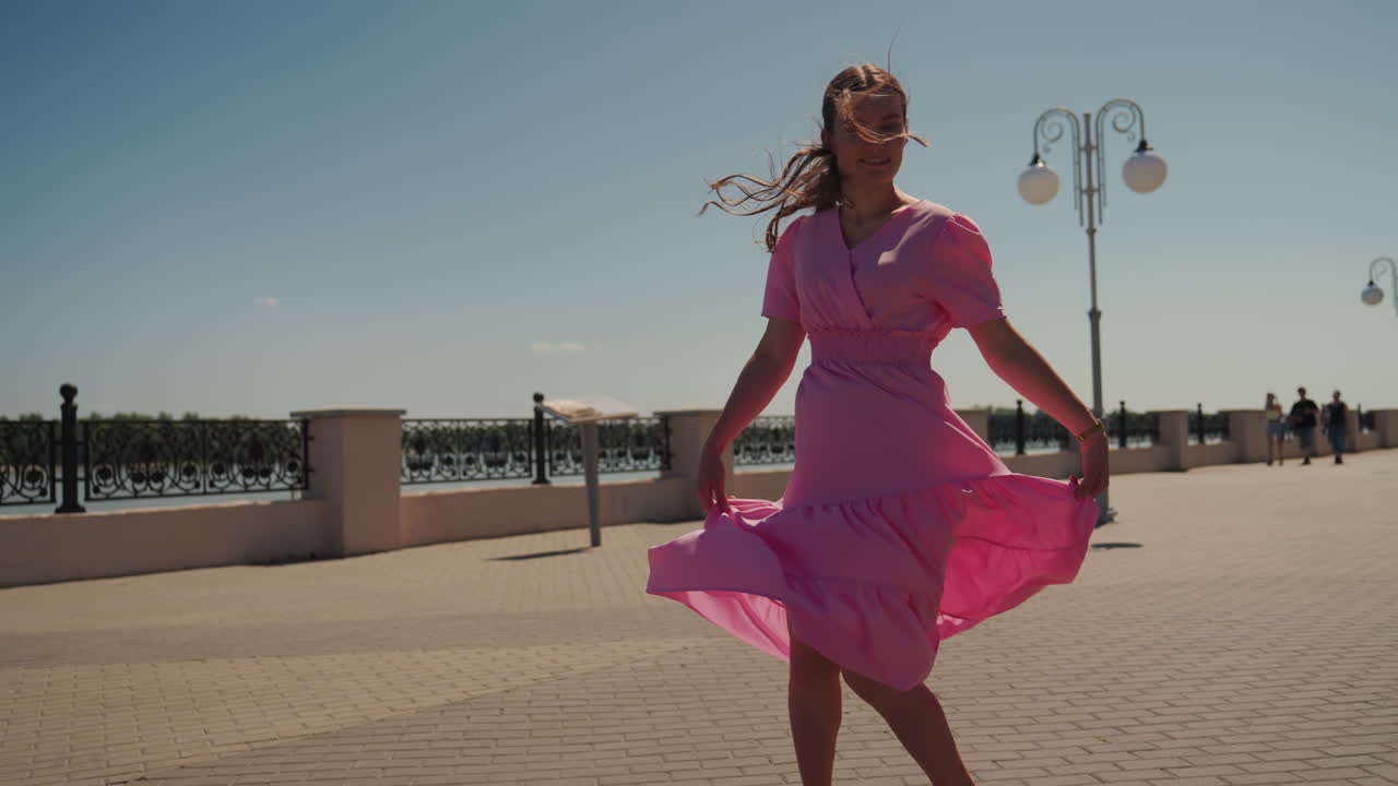 giovane donna in abito rosa passeggia sul lungomare sotto il sole splendente, turista che vaga verso le fioriere sul lungomare, modella che volteggia per la fotocamera, ballerina che si lascia accarezzare dalla brezza, estetica cinematografica estiva