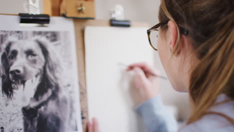 Female-Teenage-Artist-Sitting-At-Easel-Drawing-Picture-Of-Dog-From-Photograph-In-Charcoal