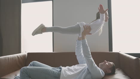 Dad-Lifting-His-Little-Daughter-Lying-On-The-Sofa-At-Home