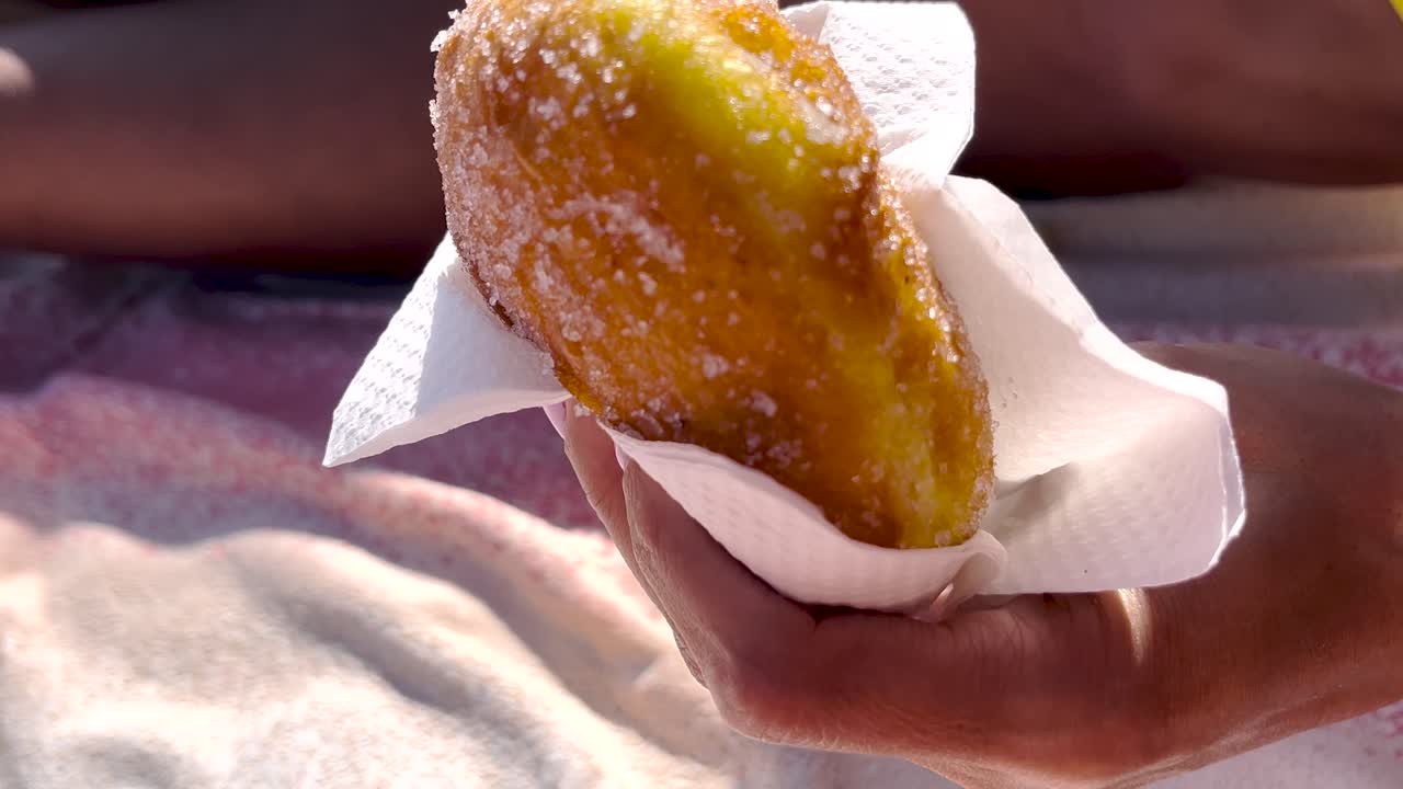 Hand holding a malasada donut with sugar at a beach food stand, close-up of fast food in colorful wrapper, yellow bikini