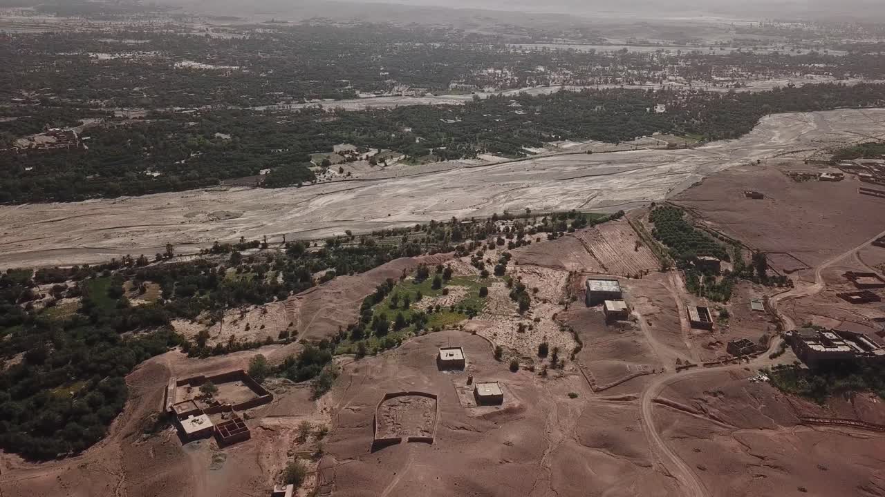 Ampia ripresa aerea del letto asciutto e sabbioso di un fiume (wadi) che separa una lussureggiante oasi verde da un terreno rurale arido. Il filmato mostra piccole strutture sparse e recinti, evidenziando la vita ai margini del deserto.