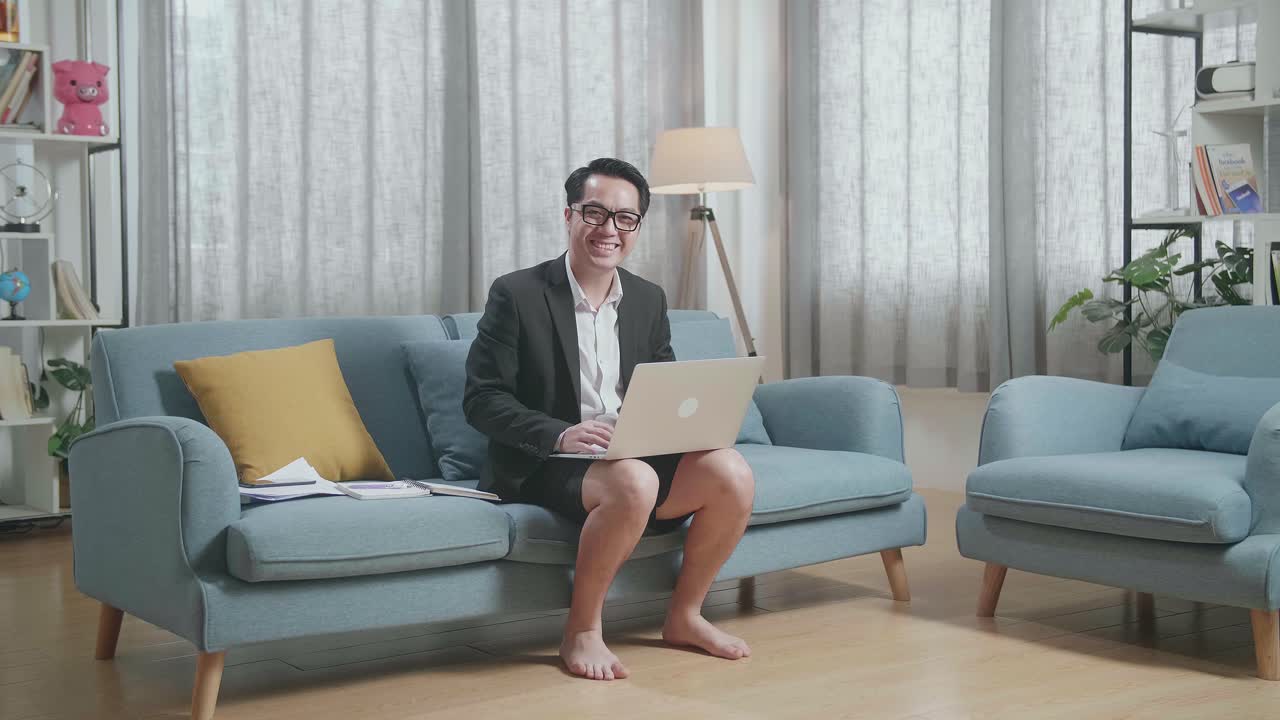 자켓과 쇼트를 입은 아시아 사업가가가 집에서 노트북을 가지고 일하면서 카메라를 보고 미소 짓고 있습니다.