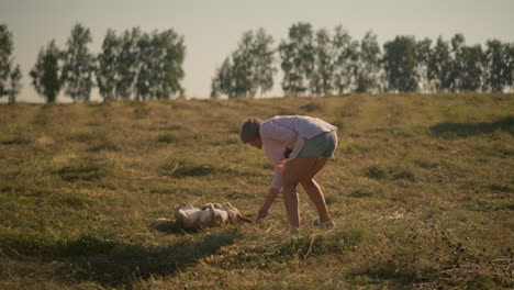 instructor de animales con camisa rosa y pantalones cortos entrenando a un perro para que se vuelva en tierras de cultivo, el perro sigue con entusiasmo la instrucción con la lengua fuera, un día soleado y brillante, con árboles lejanos en el fondo