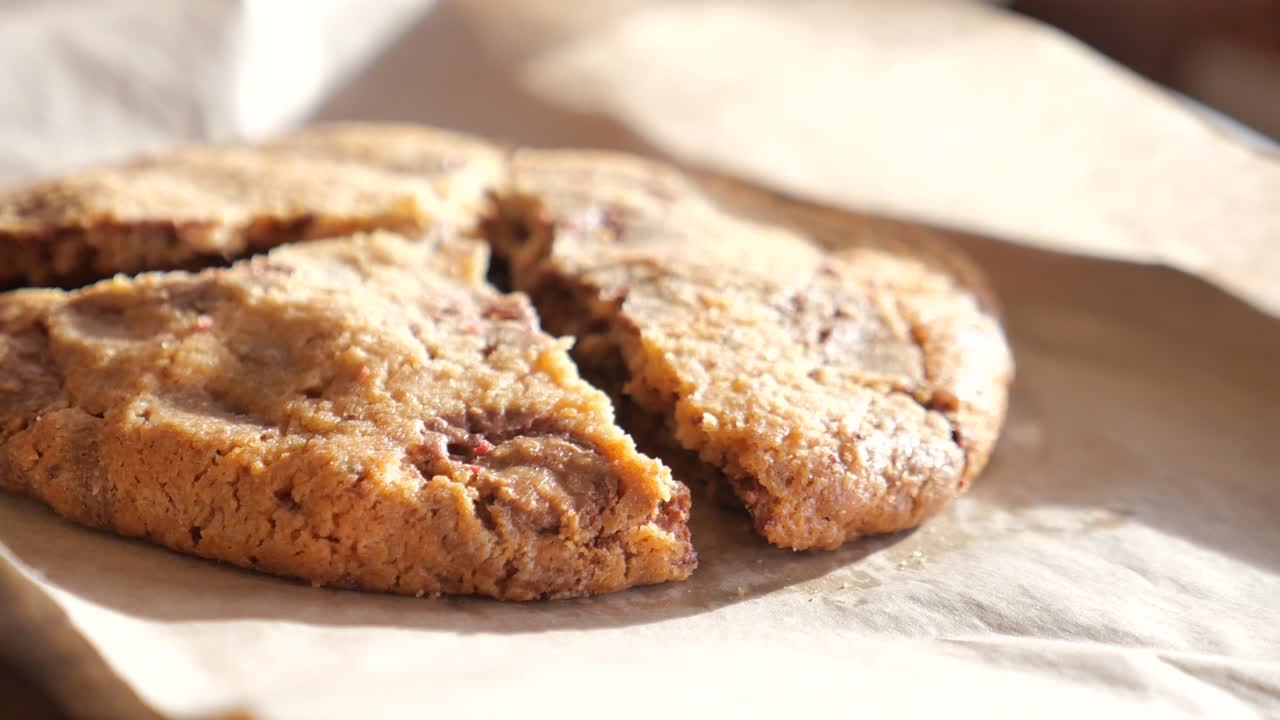 Close-up of a large chocolate chip cookie, freshly baked and split into three pieces on brown paper. The cookie is warm, soft, and golden-brown, with lots of visible chocolate chips.