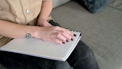 Die-Kamera-Konzentriert-Sich-Auf-Die-Hand-Einer-Frau,-Die-Ein-Braille-Buch-Liest,-Das-Zu-Hause-Auf-Dem-Sofa-Sitzt