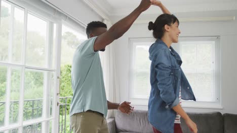 Happy-diverse-couple-holding-hands-and-dancing-in-living-room