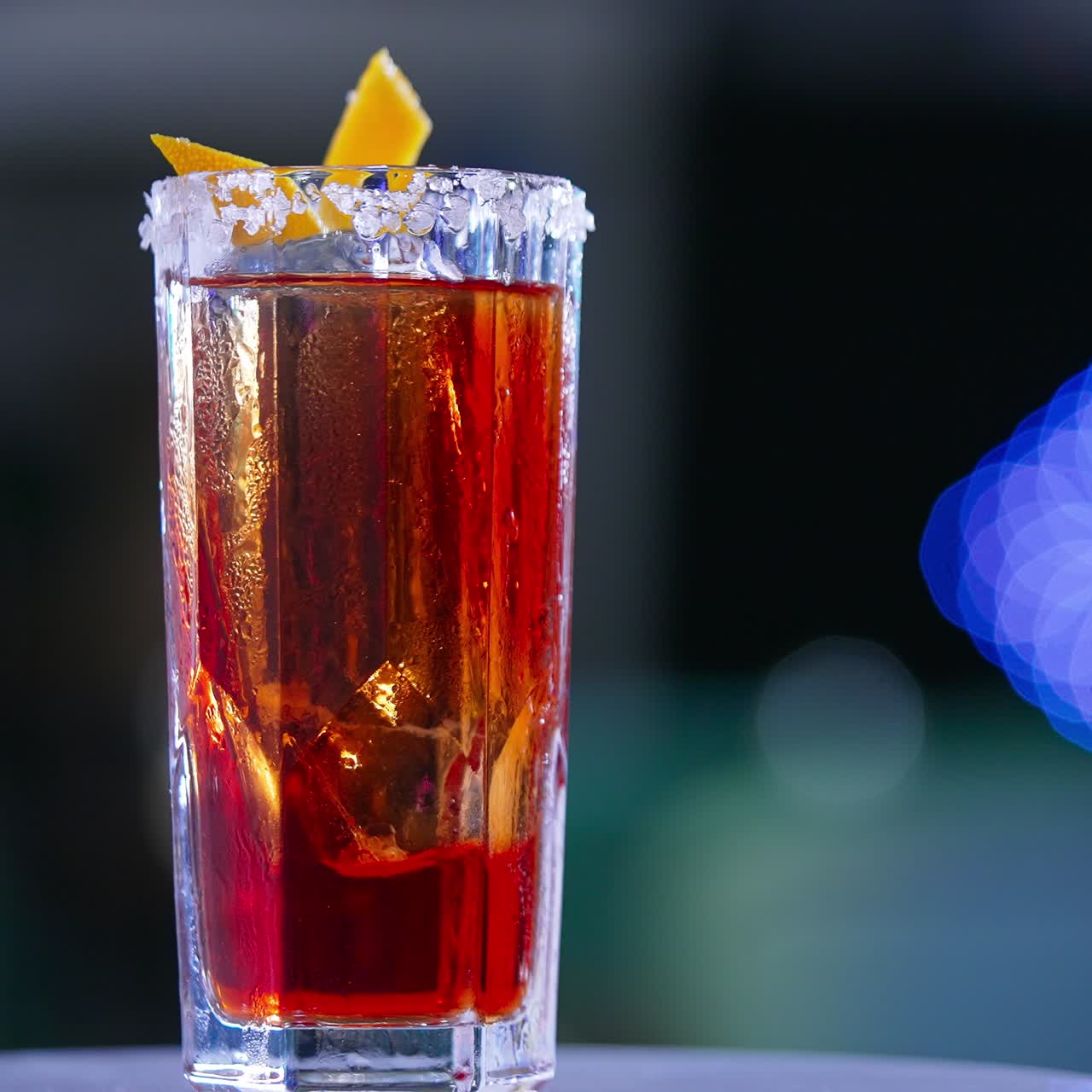 Misted long glass with red drink. Rotating table with cocktail decorated with orange peel. Close up. Blurred backdrop