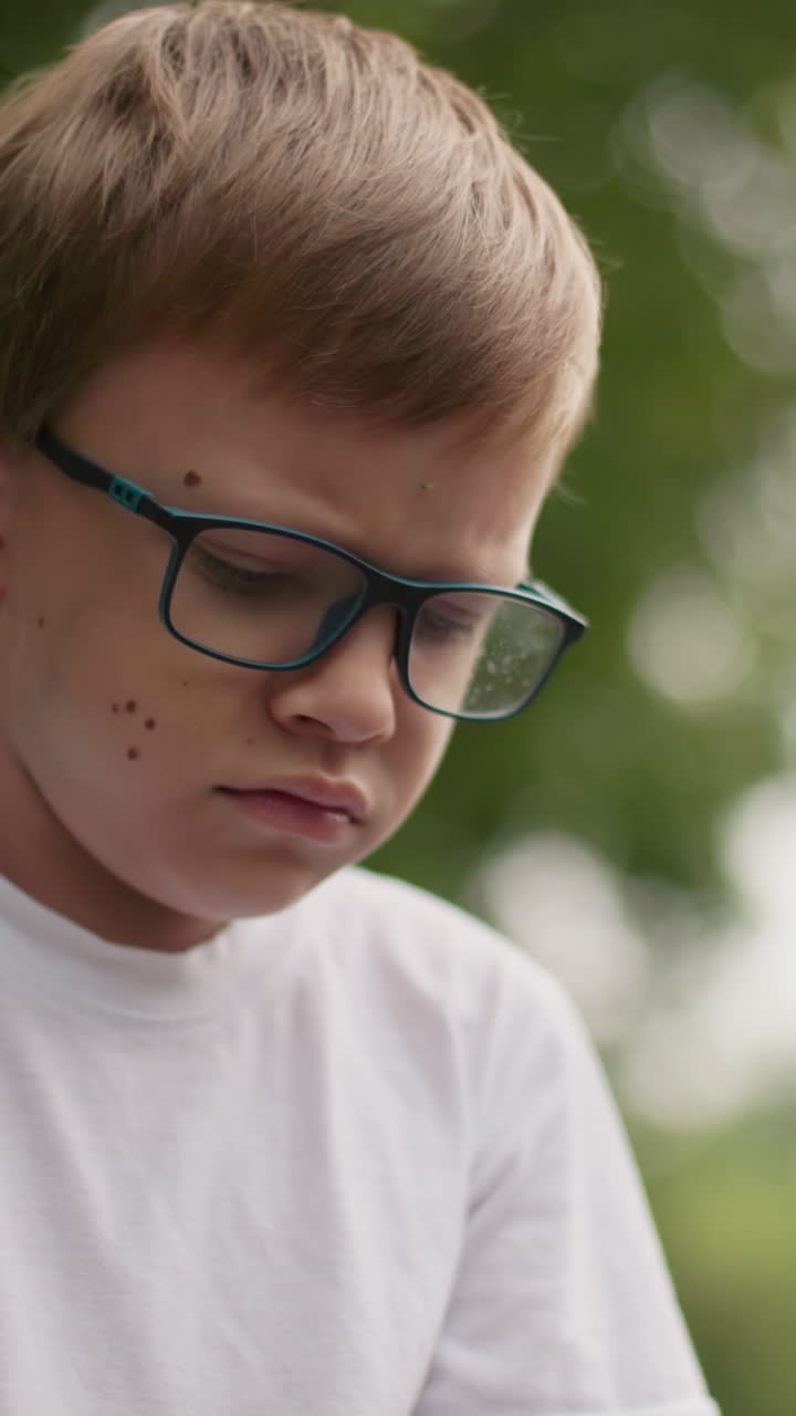 un niño con gafas parece abatido y pensativo, frunciendo el ceño ligeramente mientras mira hacia abajo, el fondo está borroso con vegetación