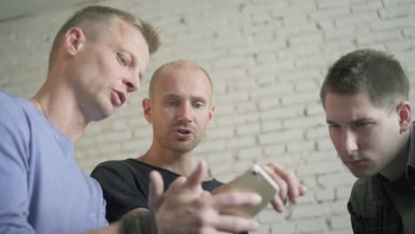 group of three buisenessmen using phone in the office while drinking coffee. males activily discussing smartphone app