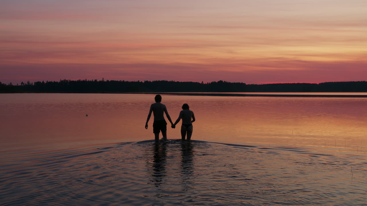 Static Shot of Silhouetted Romantic Couple Holding Hands Walking In Water to go for a Swim in Beautiful Summer Sunset Evening in Northern Sweden, Intimate Travel Adventure in Midnight Sun