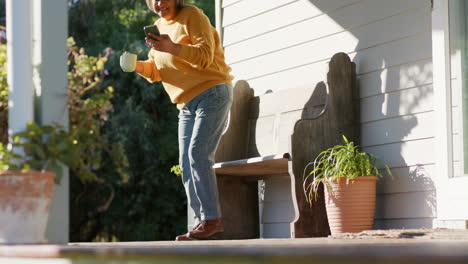 Happy-senior-biracial-woman-using-smartphone-and-drinking-coffee-on-terrace,-slow-motion,-copy-space