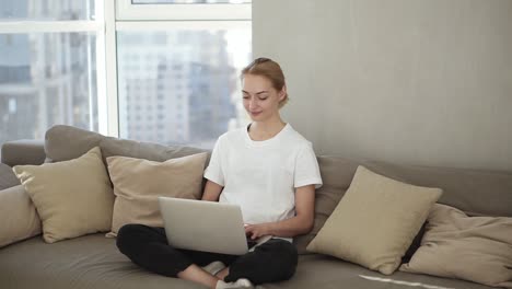 Smiling-woman-freelancer-sitting-on-couch-in-domestic-clothes,-freelance-from-home,-typing-email-on-laptop.-Focused-blonde-girl-using-computer-while-sitting-relaxfully-on-a-couch