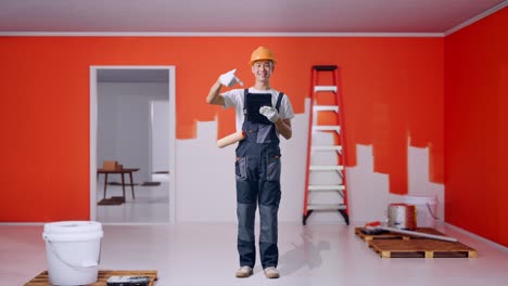 full body of asian man painter wearing safety helmet smiling and pointing to a tablet in his hand while standing with painting wall in room
