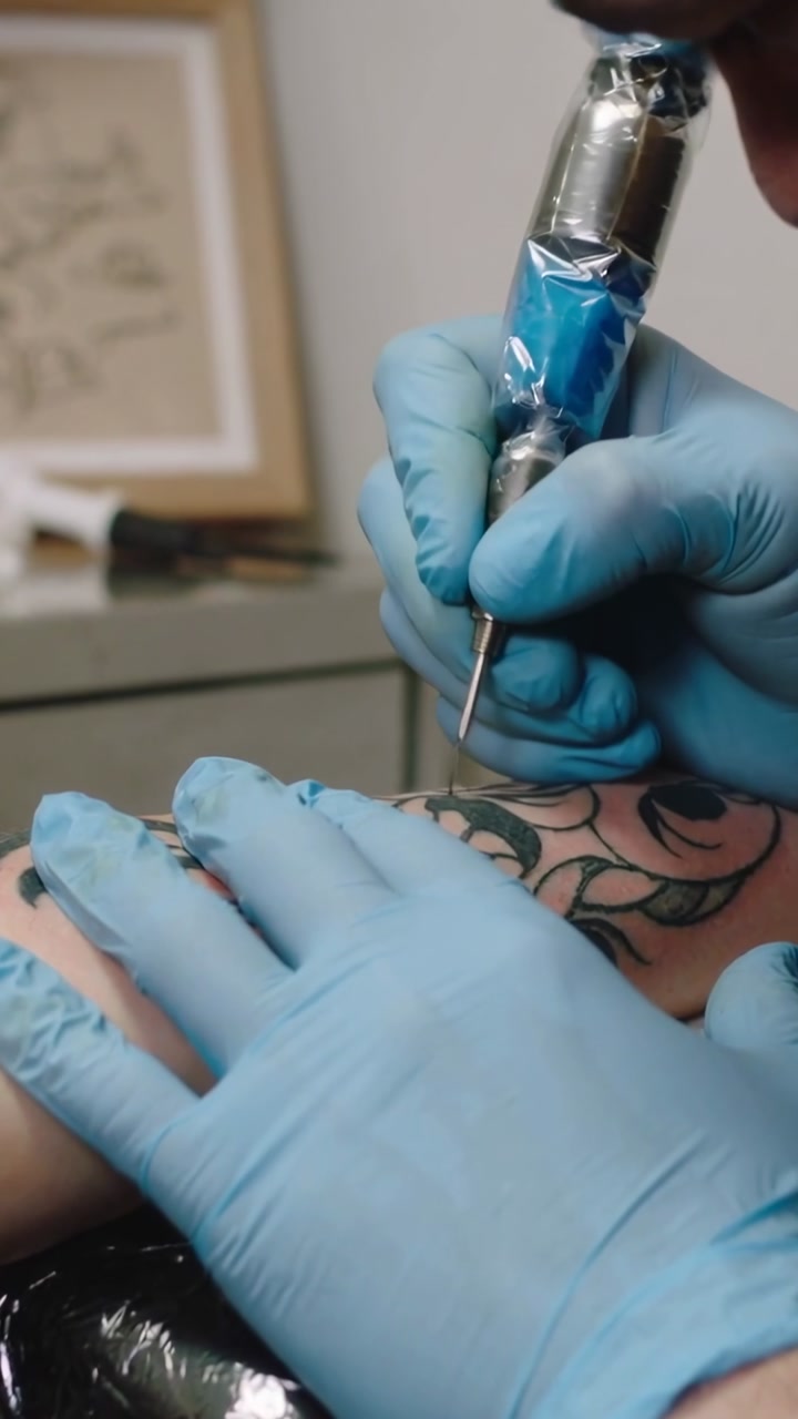 Vertical video: Needle touch urging blue-gloved artist holding arm and inking floral tattoo in shop