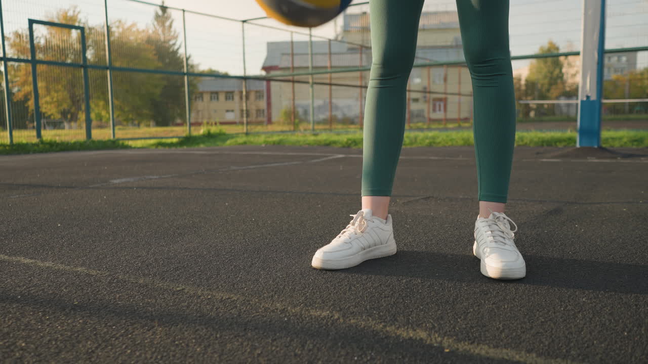 vista de cerca de la pierna de una mujer atlética rebotando voleibol al aire libre en la cancha, el aro de baloncesto y un edificio son visibles en el fondo, mostrando recreación activa en un entorno urbano vibrante