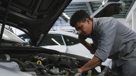 mecánico de automóviles afroamericano mirando un motor de automóvil abierto y hablando en un teléfono inteligente