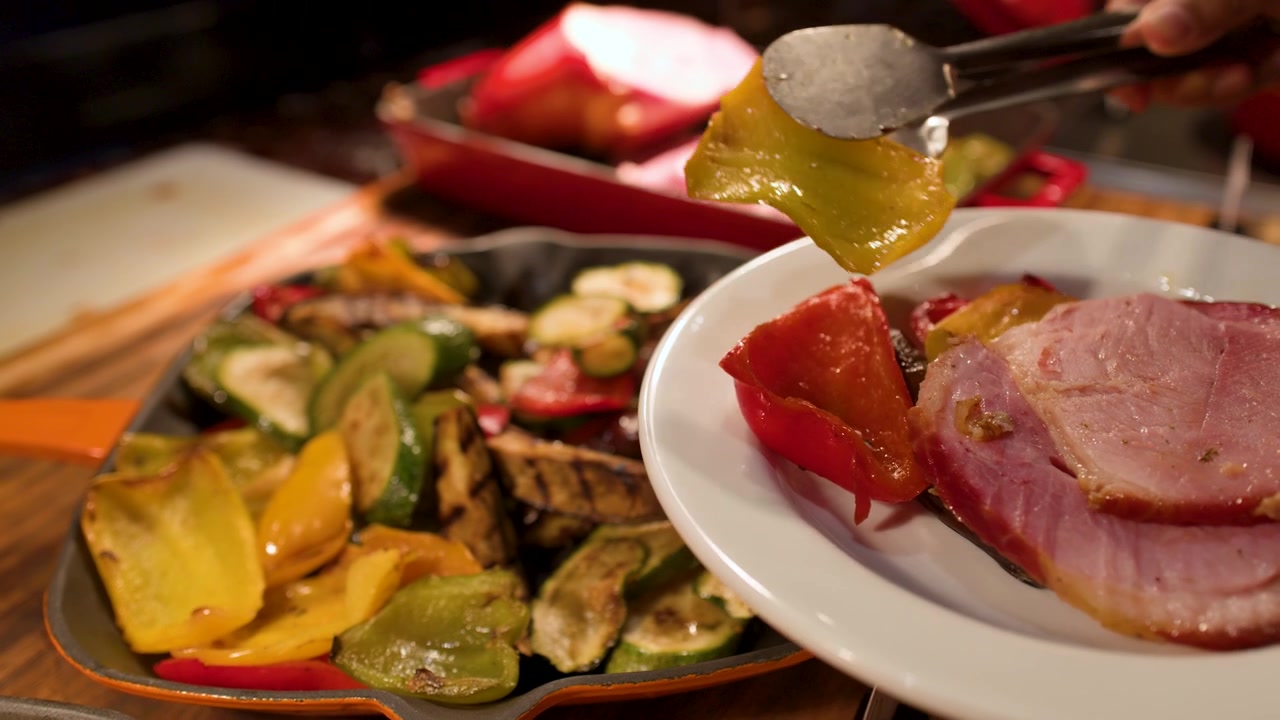 Una persona usa pinzas metálicas para servir pimientos asados en un plato con jamón