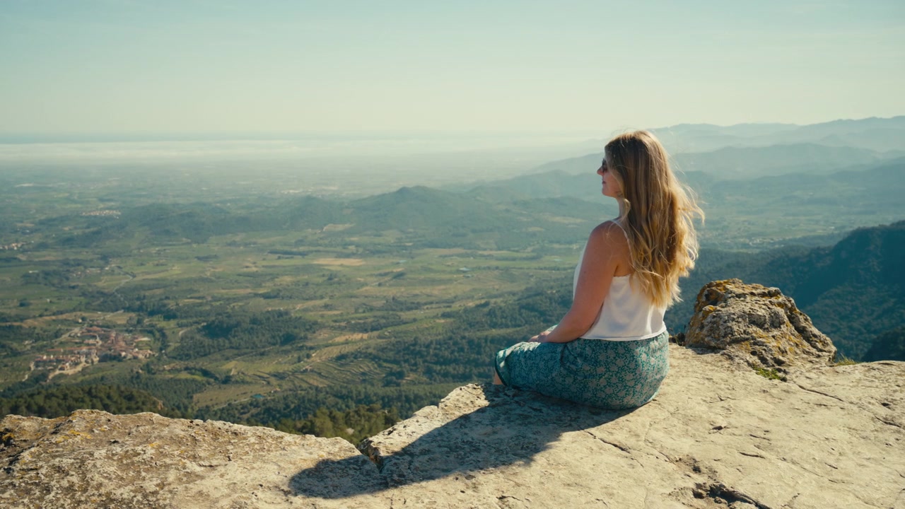 Una mujer se sienta tranquilamente en el borde de un acantilado rocoso mientras el viento mueve su cabello a cámara lenta, con vistas a un vasto valle iluminado por el sol y a las lejanas crestas montañosas en Cataluña, España