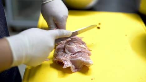 chef, con delantal de denim azul y guantes estériles blancos, destrozando cuidadosamente pollo para milanesa o milanesas, con un cuchillo afilado en una cocina en una tabla de cortar plástico amarillo