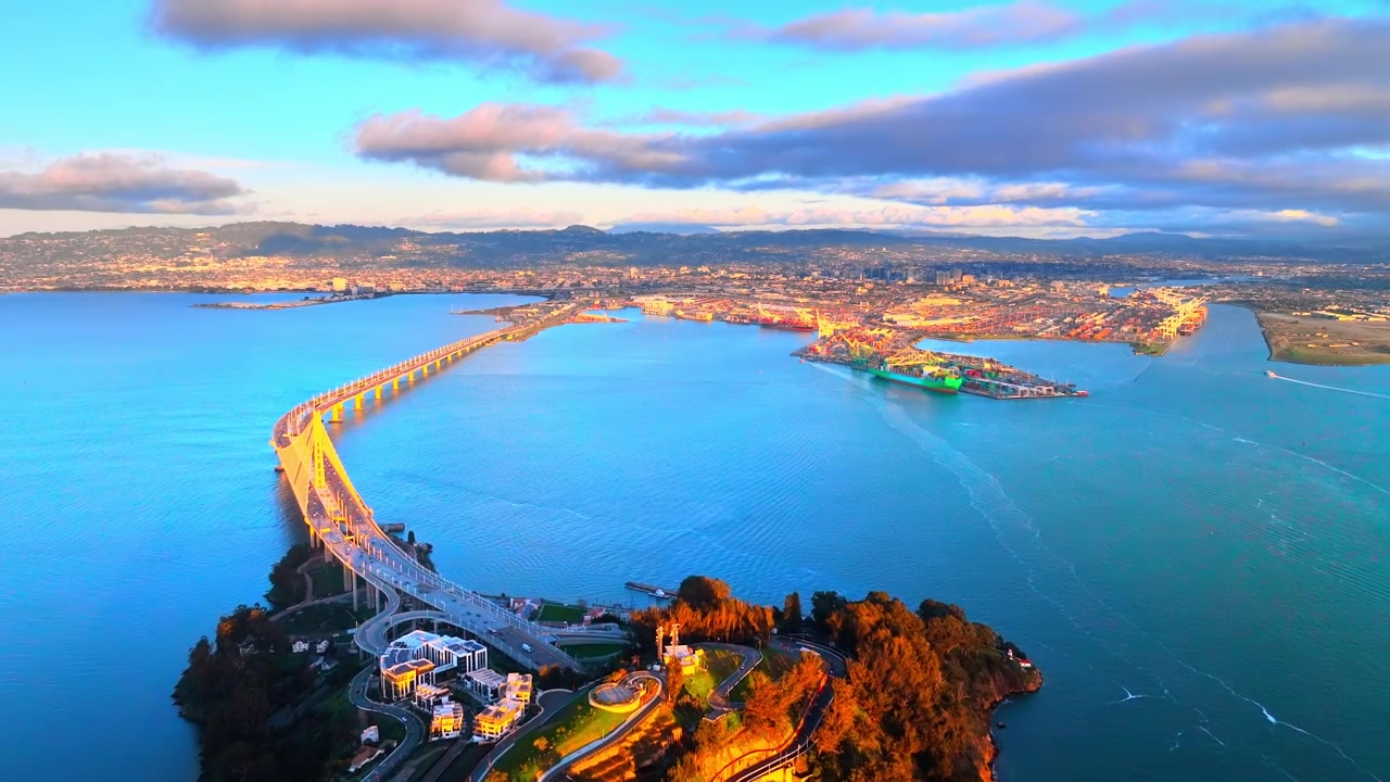 Tramonto sul Bay Bridge e sul porto di Oakland, San Francisco, California. Vista aerea di Treasure Island, gru portacontainer e navi da carico lungo la costa al tramonto.