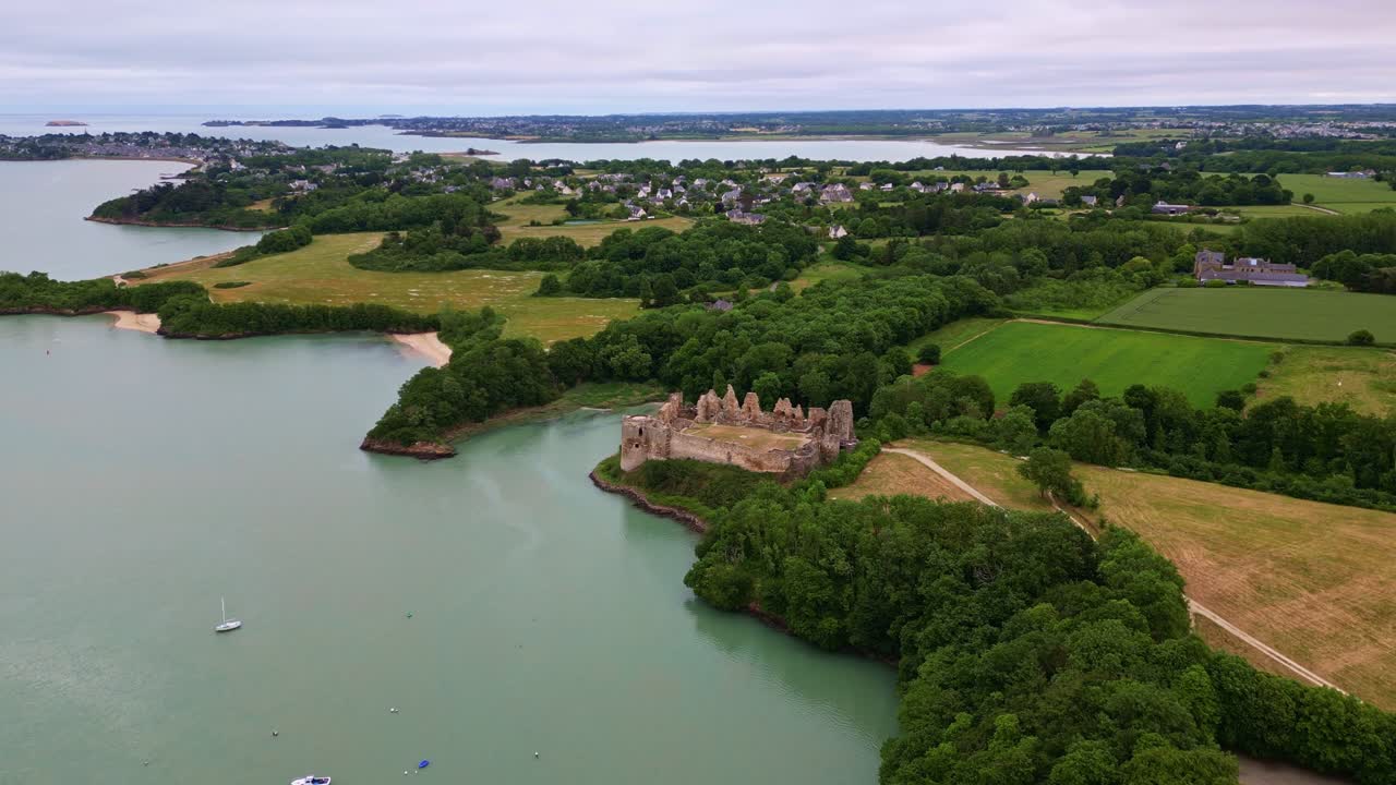 Drone flies forward toward the ruins of Guildo Castle, with surrounding forest, estuary, anchored boats and nearby villages visible. Brittany in France