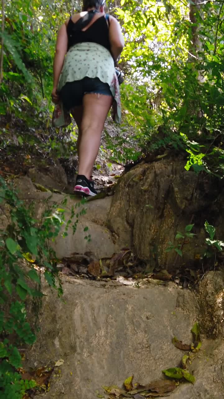 Donna che cammina in salita su un sentiero roccioso nella giungla, con vegetazione e luce solare che filtra tra gli alberi, ripresa verticale, Yelapa, Messico.