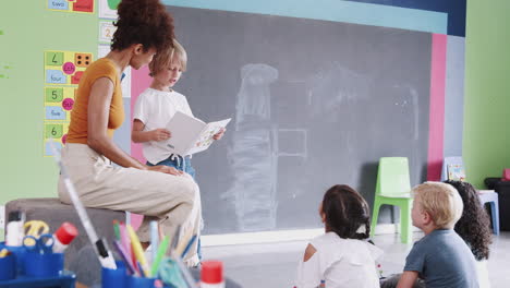 Male-Pupil-In-Elementary-School-Classroom-Reading-Book-To-Class-With-Teacher