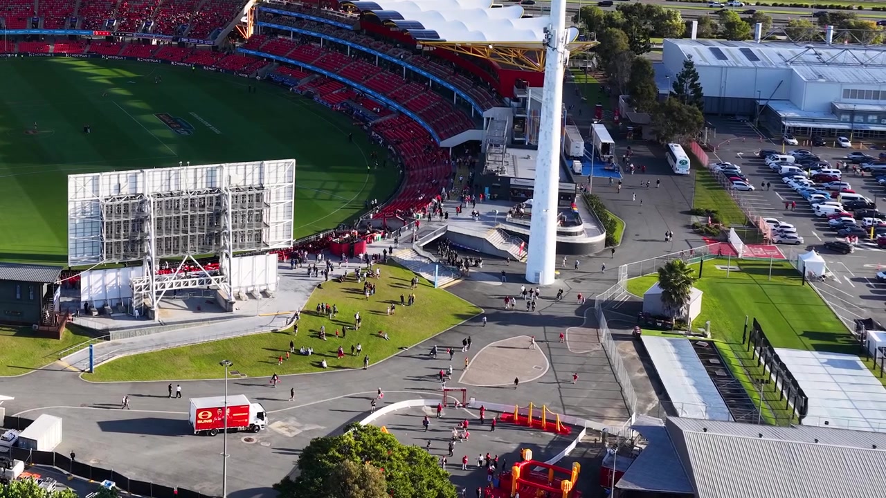 High-angle drone shot captures a large stadium complex with crowds during a sunny day.