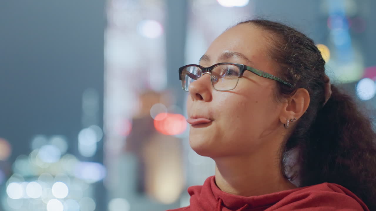 Giovane donna con gli occhiali, skyline della città di notte, viaggiatrice si ferma sul lungomare per sorseggiare una bottiglia e ammirare il panorama urbano illuminato al neon, sfondo bokeh vivace, felpa rossa con cappuccio, accenno di zaino, meraviglia urbana spontanea.