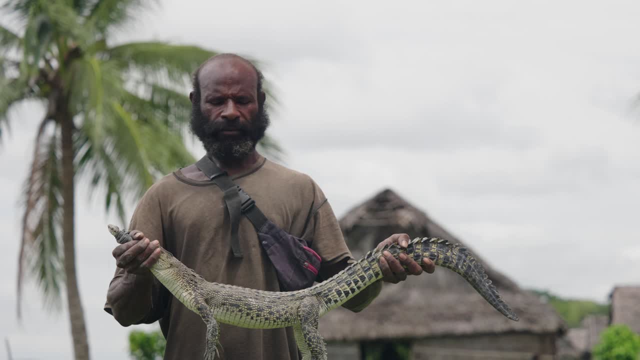 Medium Close up shot, indigenous man Holding a crocodile, blurred view hut in the background.