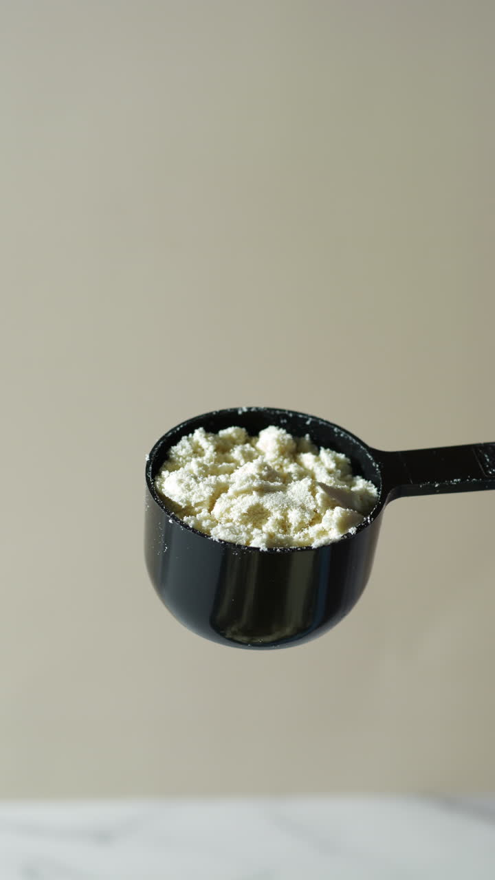 close-up of protein powder gently falling from a tilted black scoop into a white cup below, neutral beige background—highlighting the fine texture and motion of a vanilla or banana-flavored supplement