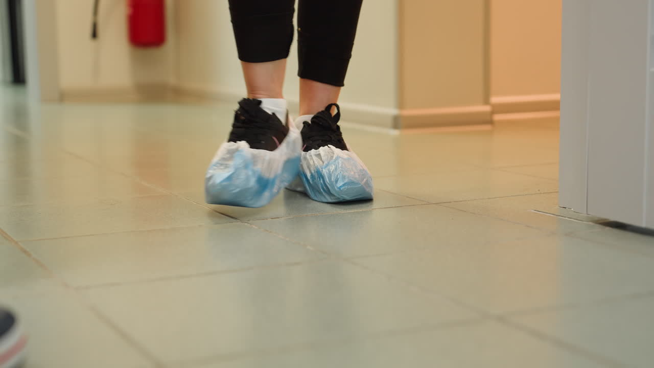 Female client wearing plastic shoe cover walks into bright white clinic room with tiled floor alongside treatment provider to cream massage chair for hygienic safety session preparation
