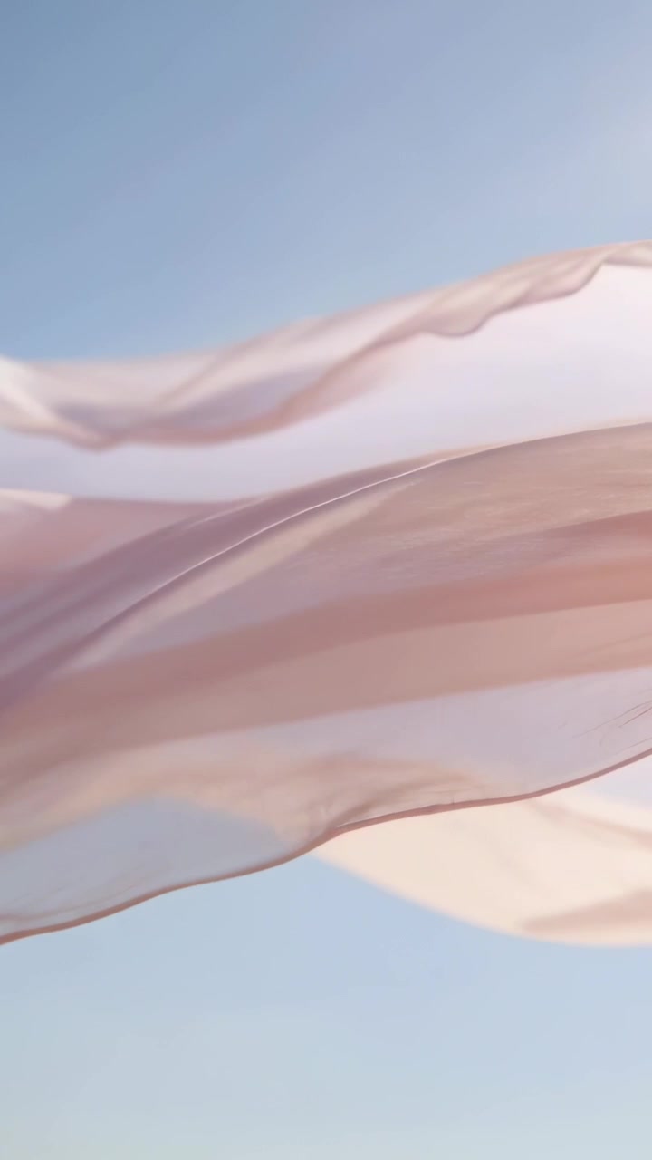 Vidéo verticale : Tissu fluide rose pâle et transparent se déplaçant sur un ciel bleu, avec une légère brise soulevant les plis.