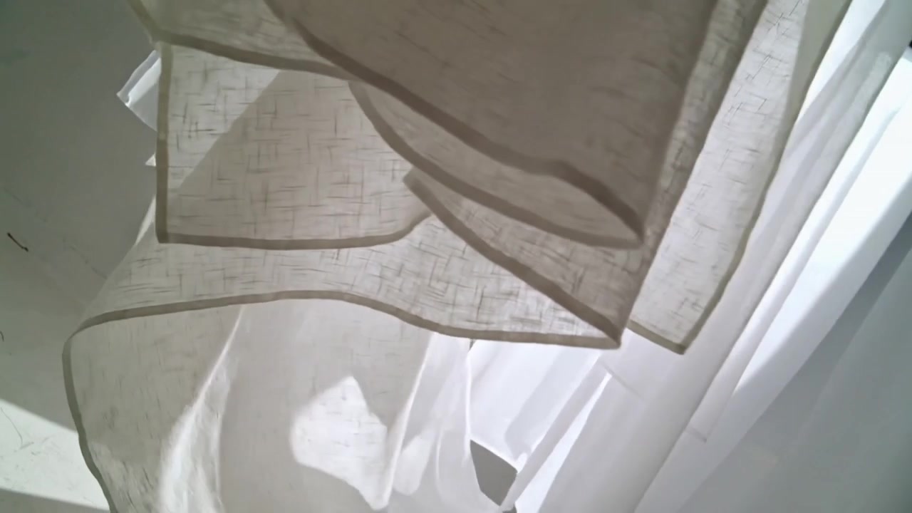 Telecamera che si sposta rivelando tende velate a strati alla finestra, mostrando pieghe fluide e un davanzale segnato.