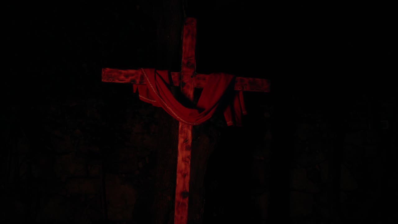 Dark scene of wooden cross with draped cloth illuminated by shifting multicolor lights creating dramatic spiritual mood