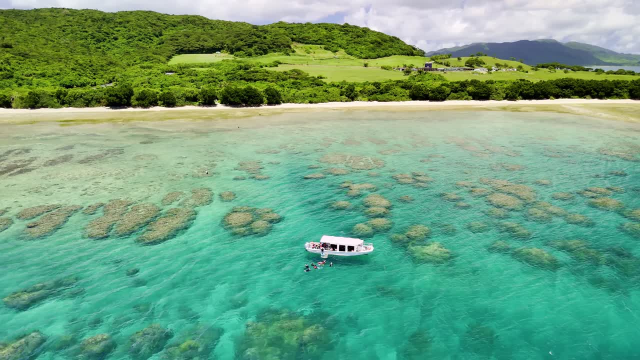 An aerial shot showcasing a boat and people snorkeling in the crystal clear turquoise water beside a lush green island. The expansive coral reef is visible just beneath the water's surface