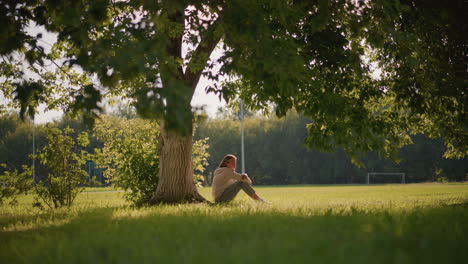 woman sits under tree in contemplative pose, arms folded over her legs, thoughtful expression on face, leaves sway gently in wind, sunlight filtering through branches, with distant goalpost