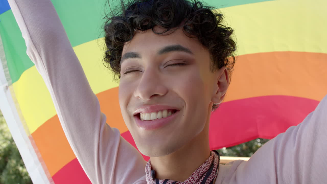 Smiling person with arms raised in front of colorful rainbow flag background