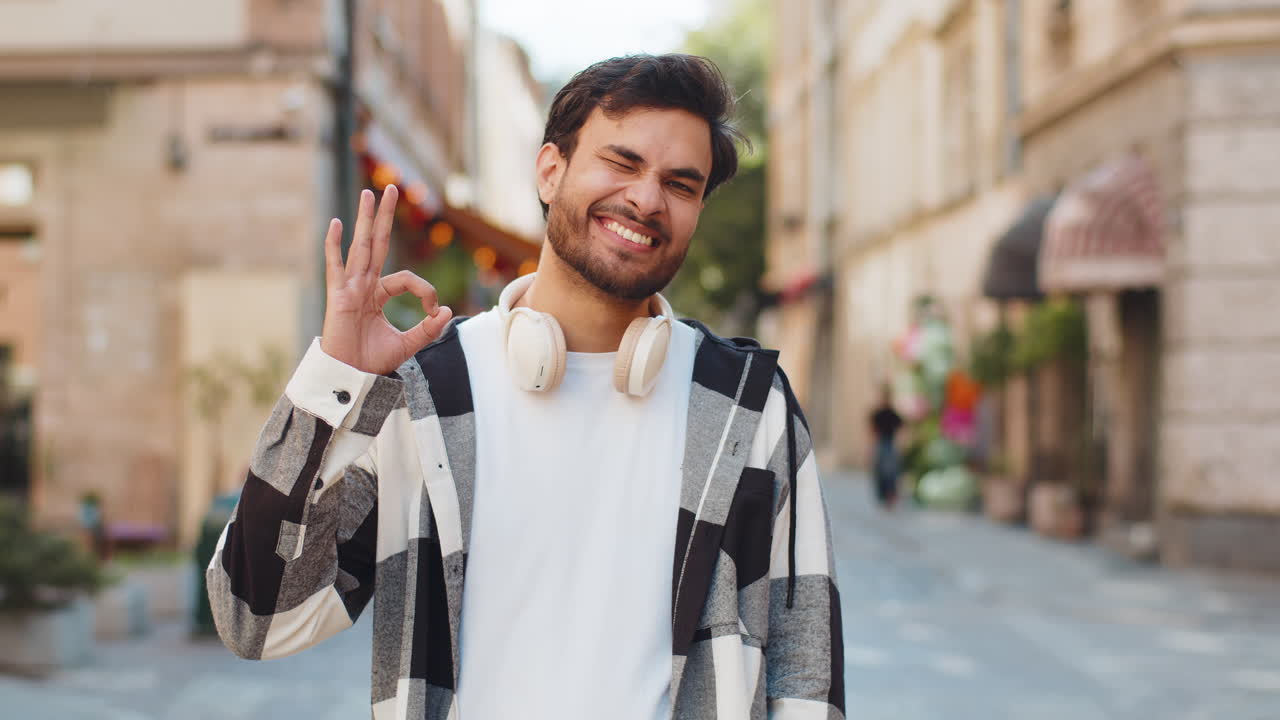 un joven indio feliz y alegre mirando con aprobación a la cámara mostrando un gesto correcto en la calle de la ciudad