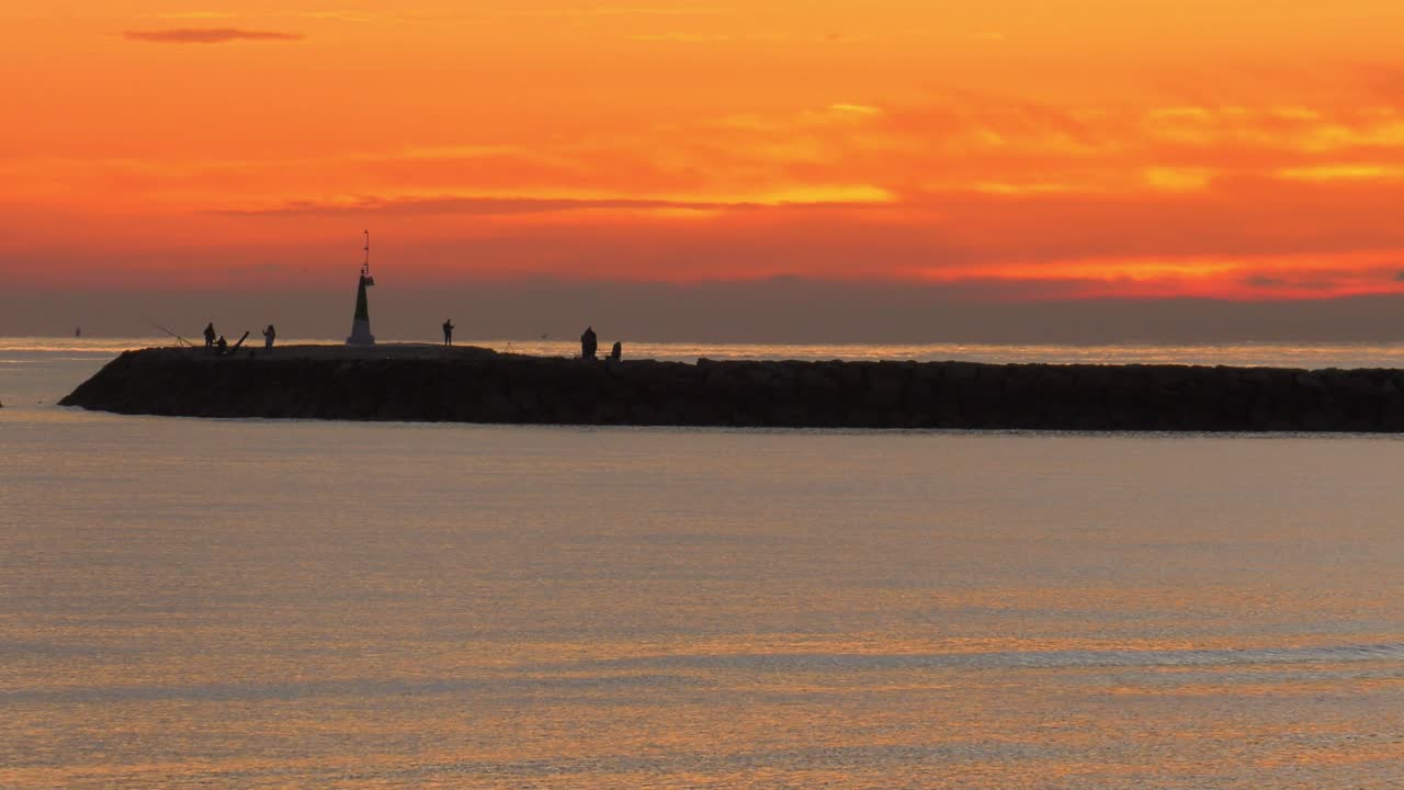 People fishing on sea wall at dawn under orange skies