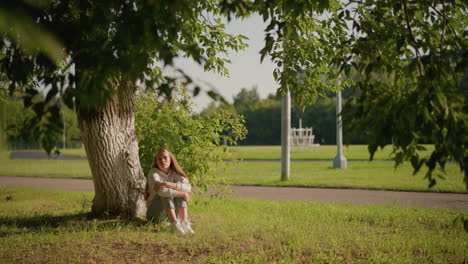 lady sits on grassy ground under tree with thoughtful expression, arms folded over legs, sunlight reflecting on her face, background shows poles and stadium