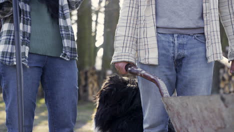 close-up view of caucasian woman petting their dog while her partner holding a wheelbarrow in the countryside