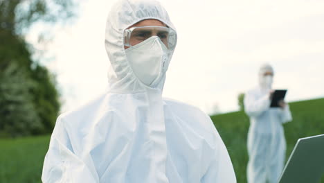 close-up view of caucasian researcher man in protective suit using laptop while doing pest control in the green field