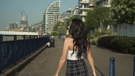 attractive and playful latina woman with black wavy hair walking on the banks of the thames river in london, looking at the camera, doing follow me gesture, happy with a beautiful smile