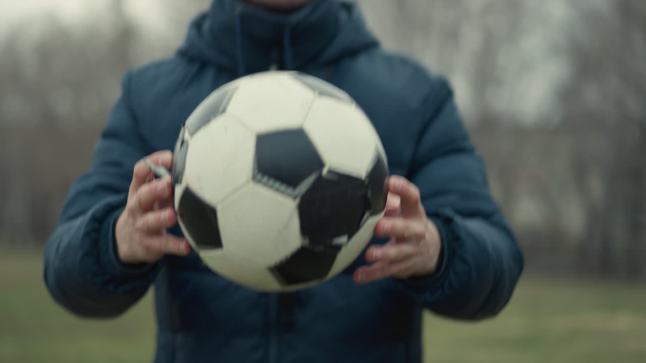 Blurred view of a coach dropping a soccer ball on a grassy field in a stadium as two people begin to play, set against a background view of buildings and trees