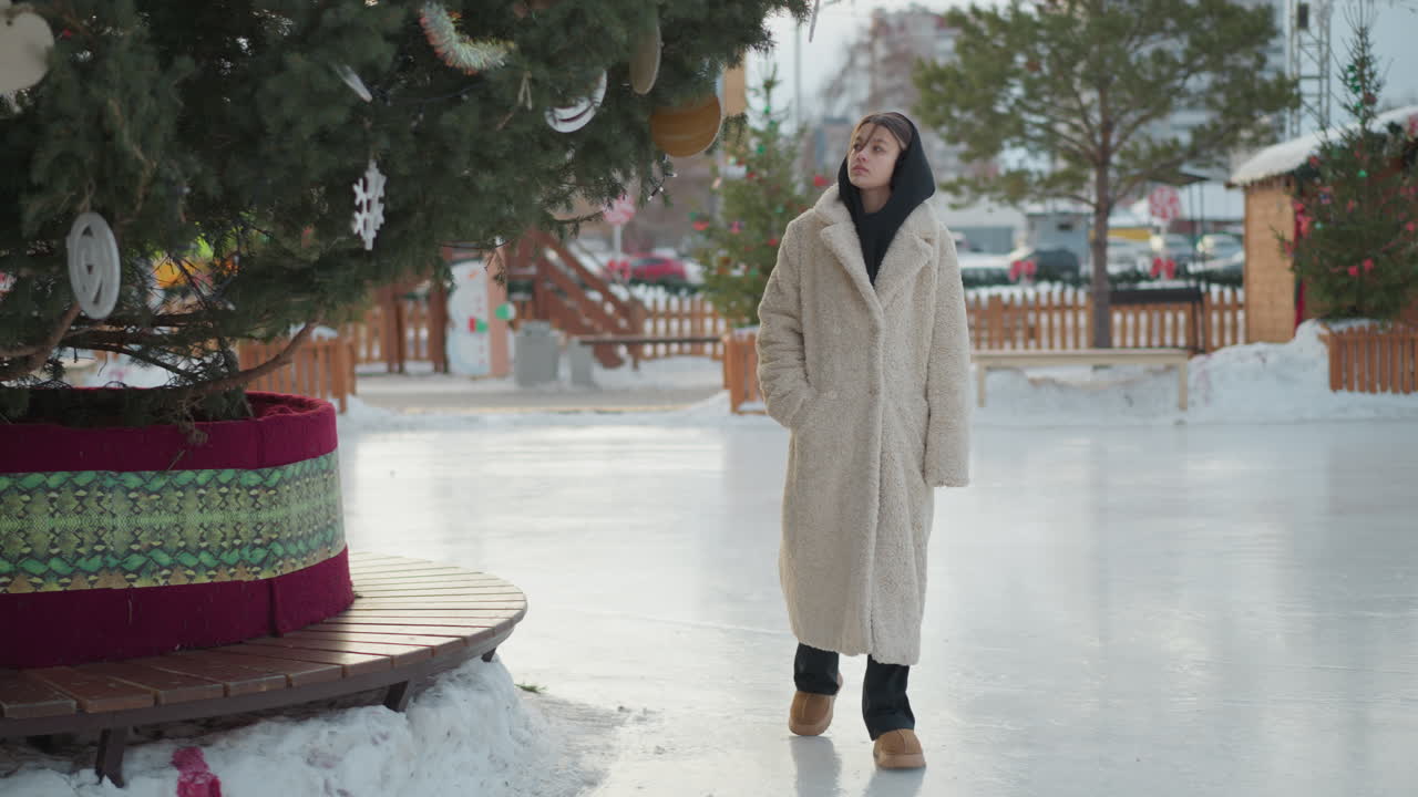 joven con una cálida chaqueta de invierno beige camina alrededor de un árbol bellamente decorado en un camino nevado, sus manos metidas en los bolsillos, mira el entorno festivo mientras los coches se mueven en el fondo
