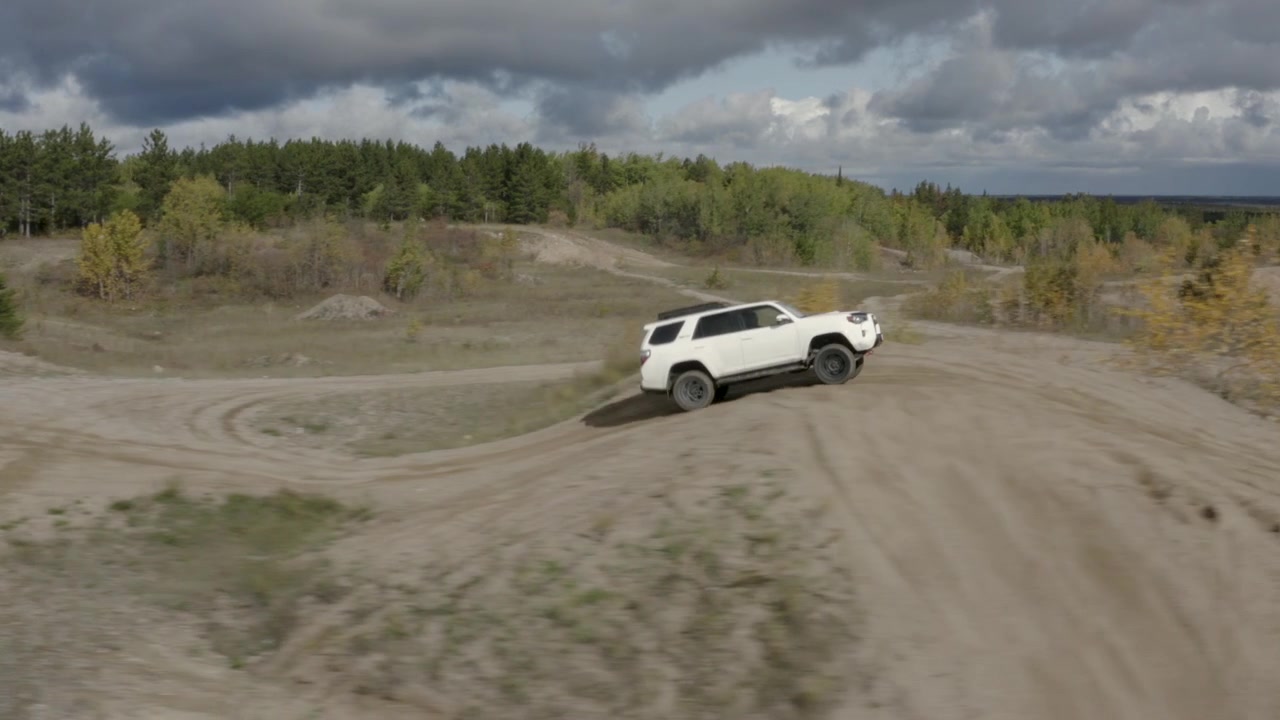 Ujęcie z lotu ptaka śledzące biały samochód omijający małe wzgórze w lesie, Szlak North Star, Manitoba, Kanada. Pochmurne niebo. Materiał 4K z drona śledzący z boku.